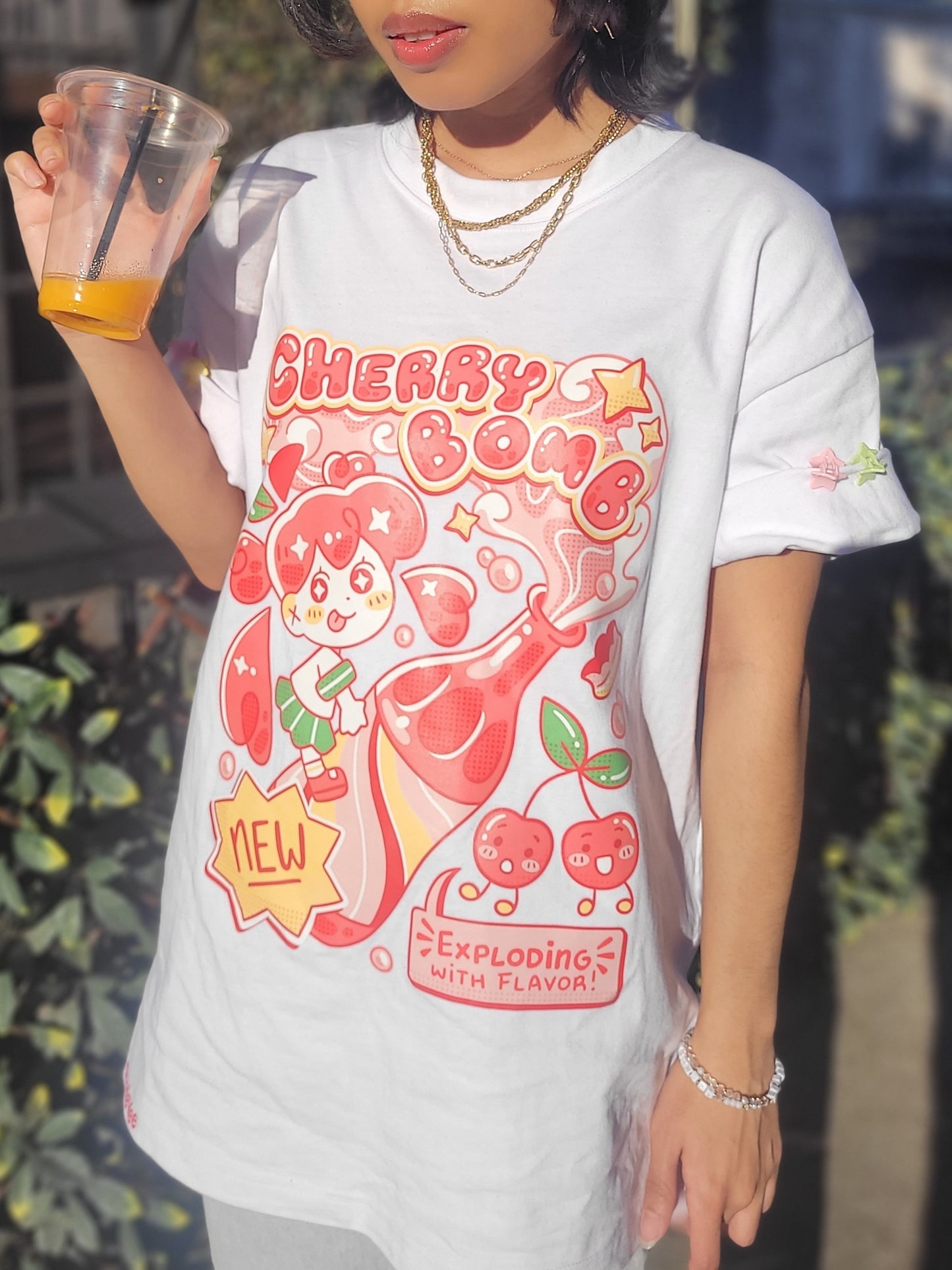 Person wearing a white t-shirt with a colorful graphic design, holding a cup outdoors.