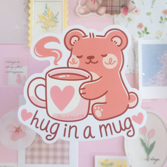 Sticker of pink bear hugging a coffee mug with the words hug in a mug on the sticker