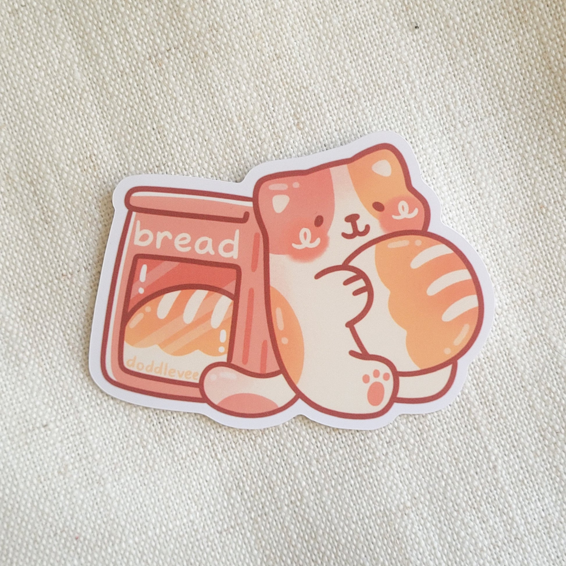 Sticker with calico cat holding bread loaf sitting next to bag with bread inside of it.