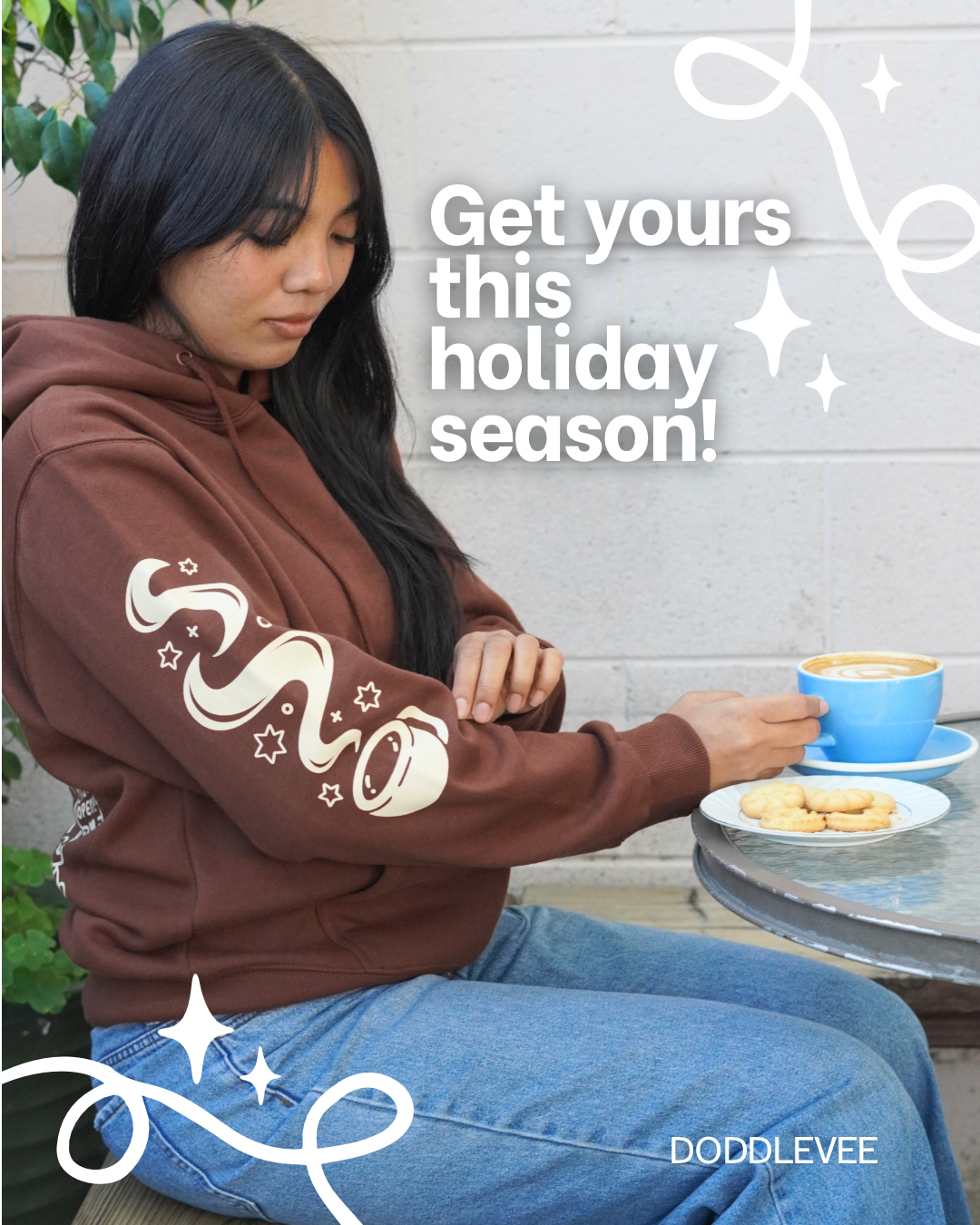 Person wearing a brown hoodie with a design, sitting outdoors with a cup of coffee and cookies.
