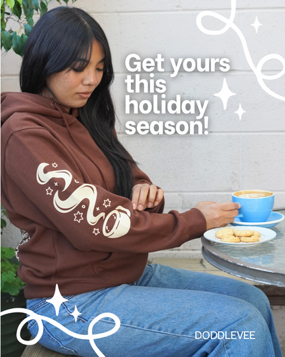Person wearing a brown hoodie with a design, sitting outdoors with a cup of coffee and cookies.